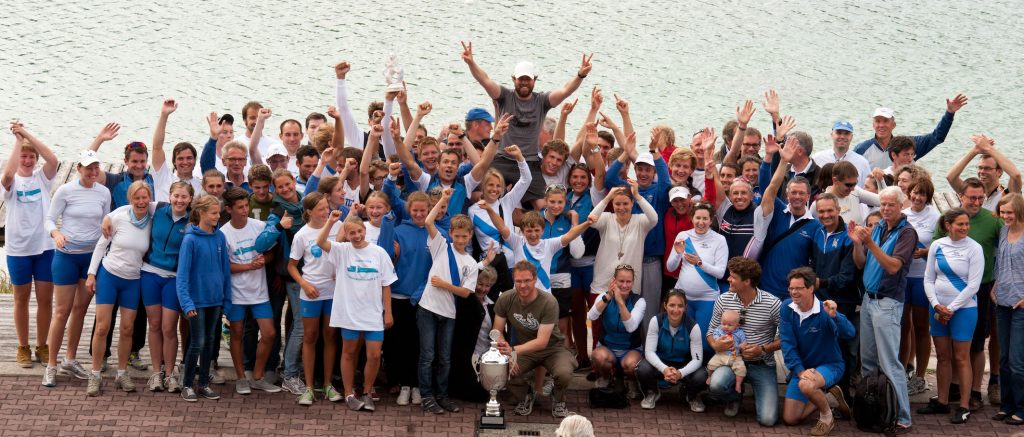 Ein Gruppenfoto von Ruderern in blau-weißer Kleidung. Einige Ruderer heben ihre Hände zum Jubel, während sie sich vor einem See befinden.