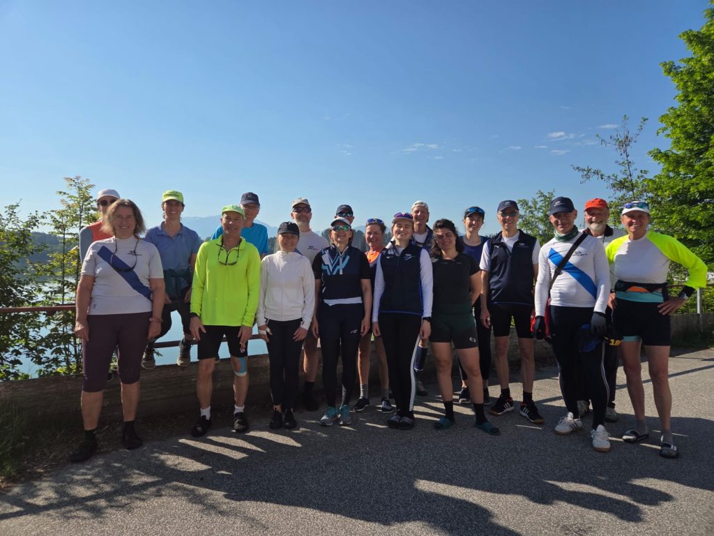 Gruppenbild lächelnder Ruderer vor See und Bergkulisse bei wolkenfreiem Himmel.